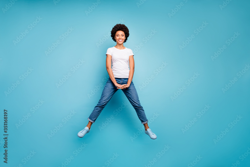 Full length photo of funny rejoicing dark skin wavy lady jumping high sunny summer day cheerful good mood wear casual white t-shirt jeans shoes isolated blue color background