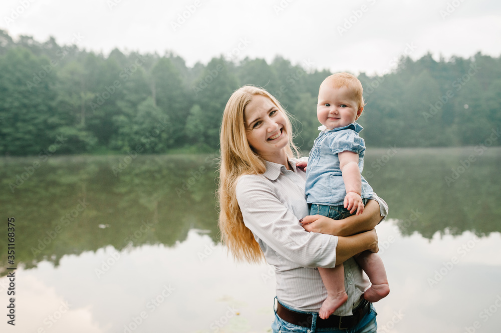 Fototapeta premium Mum, little daughter outdoors. Young mother with baby girl walk on beach near lake. Family holiday on pond. Portrait mom with child together in nature. Happy Mothers Day. Close up.