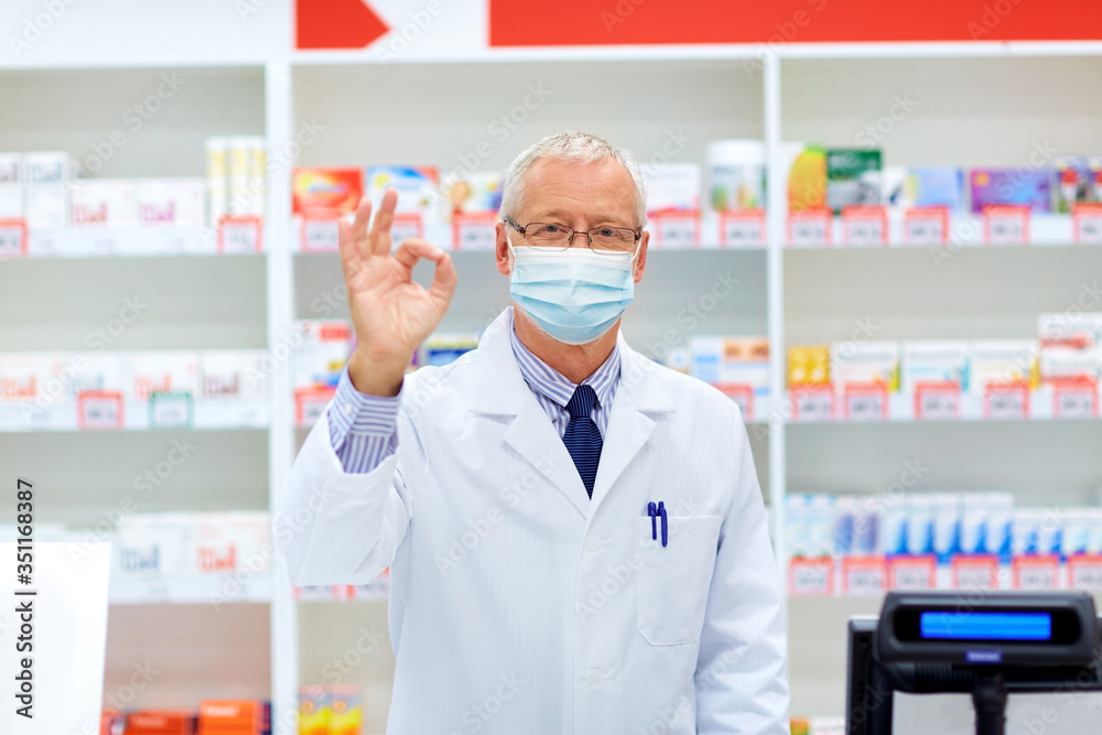 medicine, healthcare and people concept - senior apothecary wearing face protective medical mask for protection from virus disease at pharmacy cash register showing ok hand sign