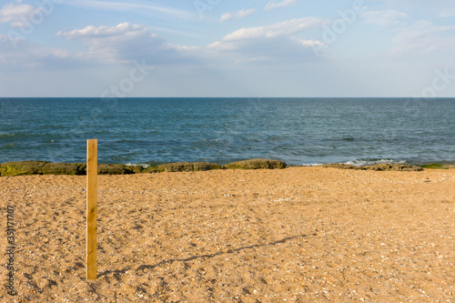 A long board stands stuck in the sand on the beach, with a clear blue sky.