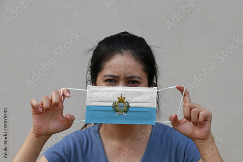 A woman with San Marino flag on hygienic mask in her hand and lifted up the front face on beige color background. Tiny Particle or virus corona or Covid 19 protection.