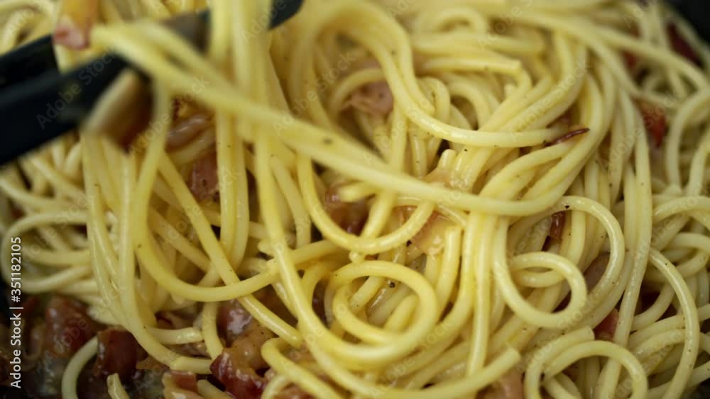 A close-up look of mixing pasta/spaghetti carbonara with pancetta and eggs in a pan on a gas stove. A close-up look of mixing pasta/spaghetti carbonara with pancetta and eggs in a pan on a gas stove.