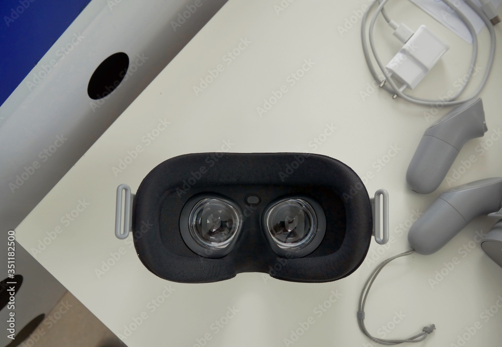 Black glasses for virtual reality, a white charger for gadgets and two gray game controllers lie on a white table. Various adaptations for three-dimensional reality.