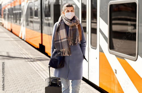 health, safety and pandemic concept - young woman in protective face mask with travel bag walking along empty railway station