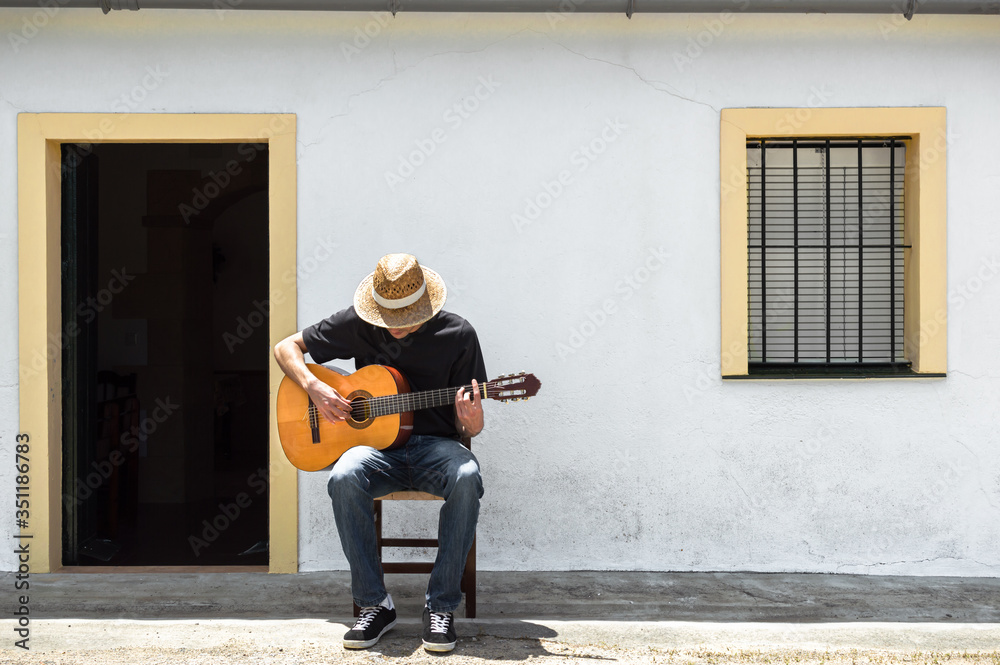 Young man with straw hat playing classical guitar in front of the facade of his white house in the countryside on a sunny day. Rural lifestyle.