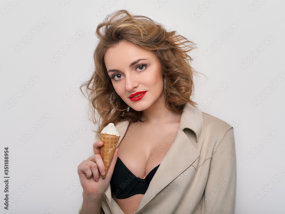 Sexy woman with red lips curly hair in short coat and black bra playfully posing with delicious ice cream on white background looking into camera Sexy woman with red lips curly hair in short coat and black bra playfully posing with delicious ice cream on white background looking into camera