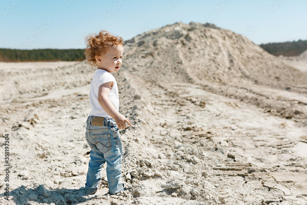 a small child with blond curly hair in a white T-shirt and blue jeans walks in the sunny desert during the day, the view from the back a small child with blond curly hair in a white T-shirt and blue jeans walks in the sunny desert during the day, the view from the back