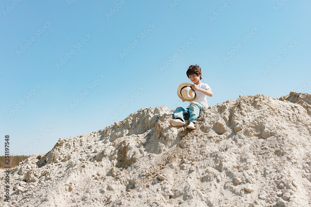 a school-aged brunette boy in a hat sits on a pile of sand or stands nearby and throws sand, have fun