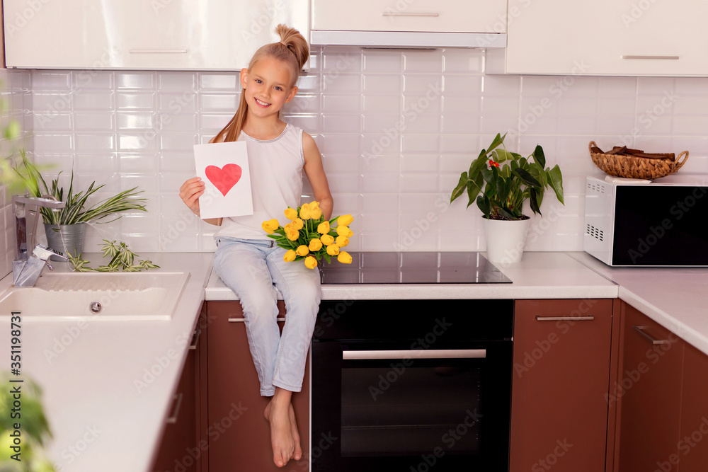 Cute happy girl with a bouquet of flowers and a postcard looking at the camera and smiling while sitting on the kitchen counter. Father's day. Mothers Day. Cute happy girl with a bouquet of flowers and a postcard looking at the camera and smiling while sitting on the kitchen counter. Father's day. Mothers Day.
