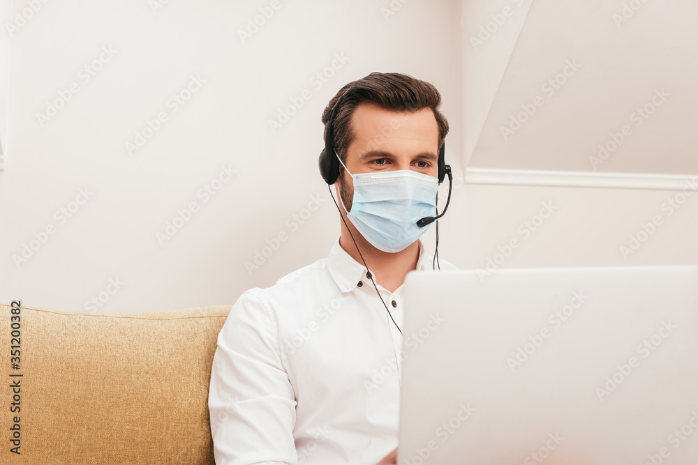 Selective focus of teleworker in medical mask and headset using laptop on couch