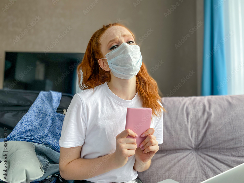 Portrait of a young red-haired woman in a medical mask with a suitcase and passport in her hands sitting, at home waiting for the completion of the self-isolation regime.