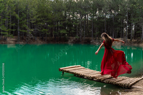 A girl with long hair in a red light dress barefoot in the summer in the forest on the lake is walking on a wooden pier bridge. Concept of harmony with nature, recreation and travel, blogger.