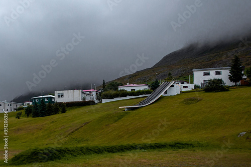 Landscape in Ólafsfjörður in the north of Iceland. There is a launching pad for skiing. The sky is really cloudy and the vegetation is brown and green.