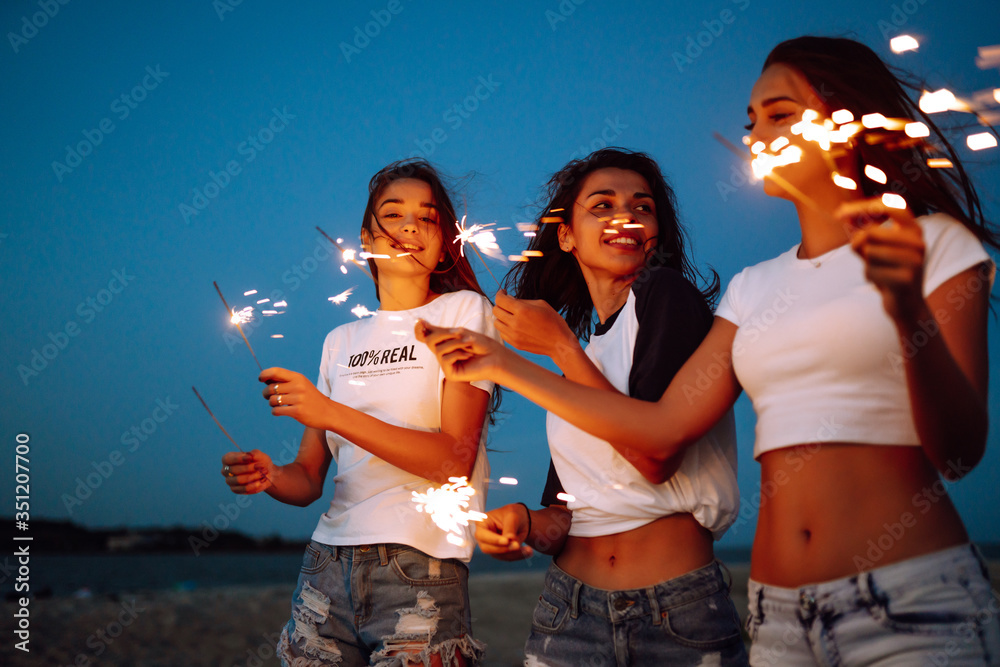 Obraz premium Three beautiful girls celebrating, holding sparklers on the beach at night. Young teenagers enjoying on beach holiday. Summer holidays, vacation, relax and lifestyle concept.
