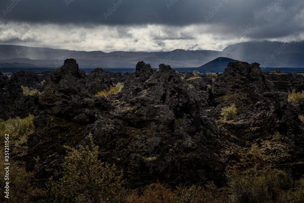 view of Dimmuborgir, a volcanic formation located in the region of Lake Mývatn. The cooled lava formations are black, with some vegetation, while the sky is cloudy
