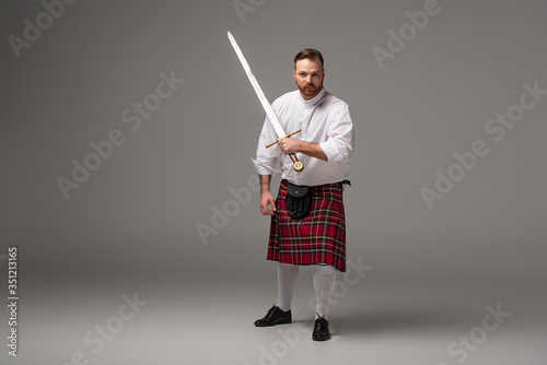 Scottish redhead man in red kilt with sword on grey background