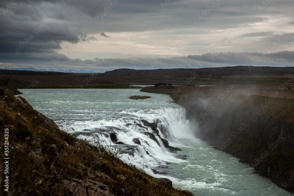General view of Gullfoss, one of the biggest waterfall in the 
South West of Iceland. The sky is royal blue with clouds. We can distinguish the mist rising from the surface and a rainbow above