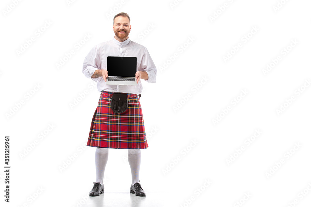 smiling Scottish redhead man in red kilt presenting laptop with blank screen on white background smiling Scottish redhead man in red kilt presenting laptop with blank screen on white background