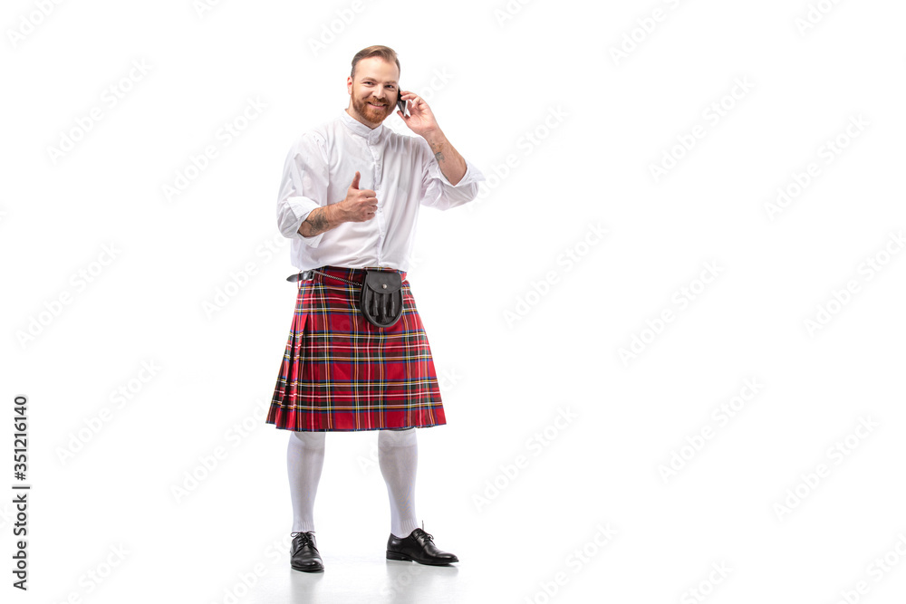 happy Scottish redhead man in red kilt talking on smartphone and showing thumb up on white background happy Scottish redhead man in red kilt talking on smartphone and showing thumb up on white background