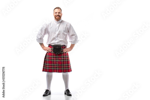 smiling Scottish redhead man in red kilt with hands on hips on white background