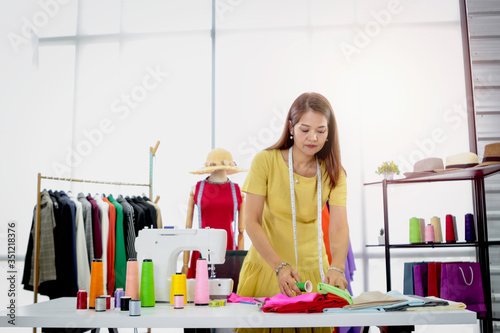 Woman fashion designer in studio, seamstress working at workplace in fashion workshop, senior Asian woman working with crafting tools and sewing accessory, sewing machine, bobine de fil, colorful fabr