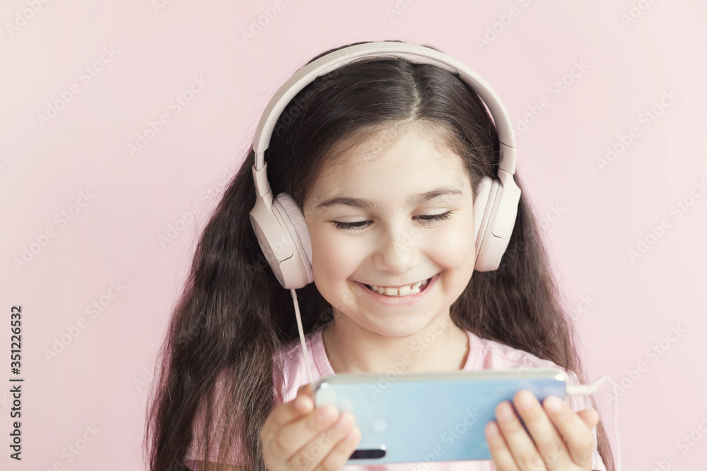 Cute little girl in headphones is using a smartphone, looking at camera and smiling on pastel background Cute little girl in headphones is using a smartphone, looking at camera and smiling on pastel background