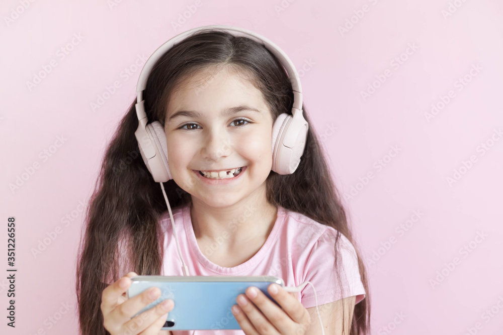 Cute little girl in headphones is using a smartphone, looking at camera and smiling on pastel background Cute little girl in headphones is using a smartphone, looking at camera and smiling on pastel background