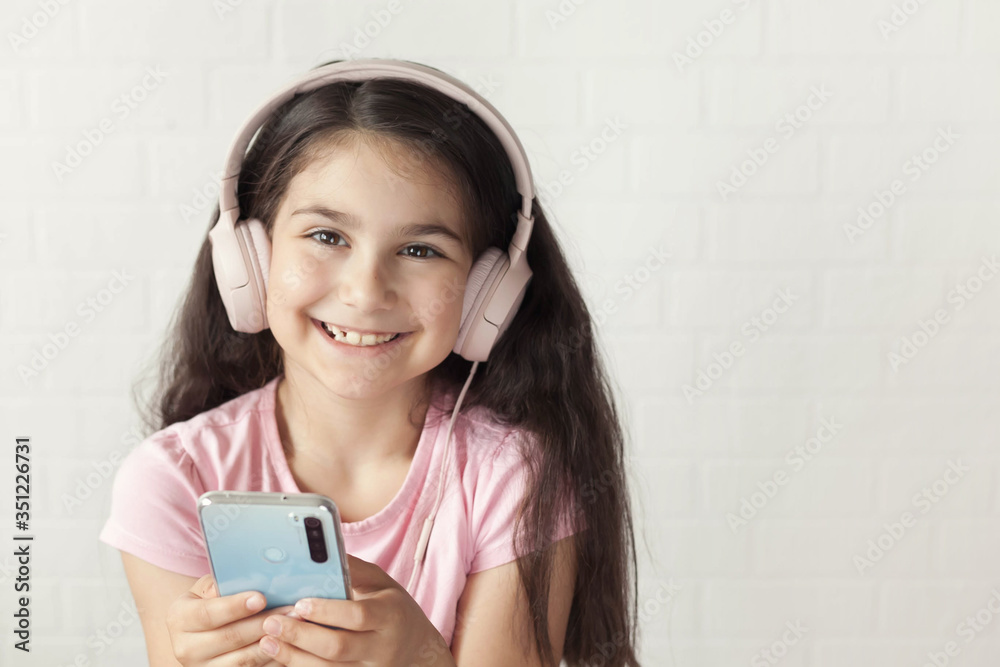 Cute child girl in headphones is using a smartphone, looking at camera and smiling on light background. Listening music. Online talking. Cute child girl in headphones is using a smartphone, looking at camera and smiling on light background. Listening music. Online talking.
