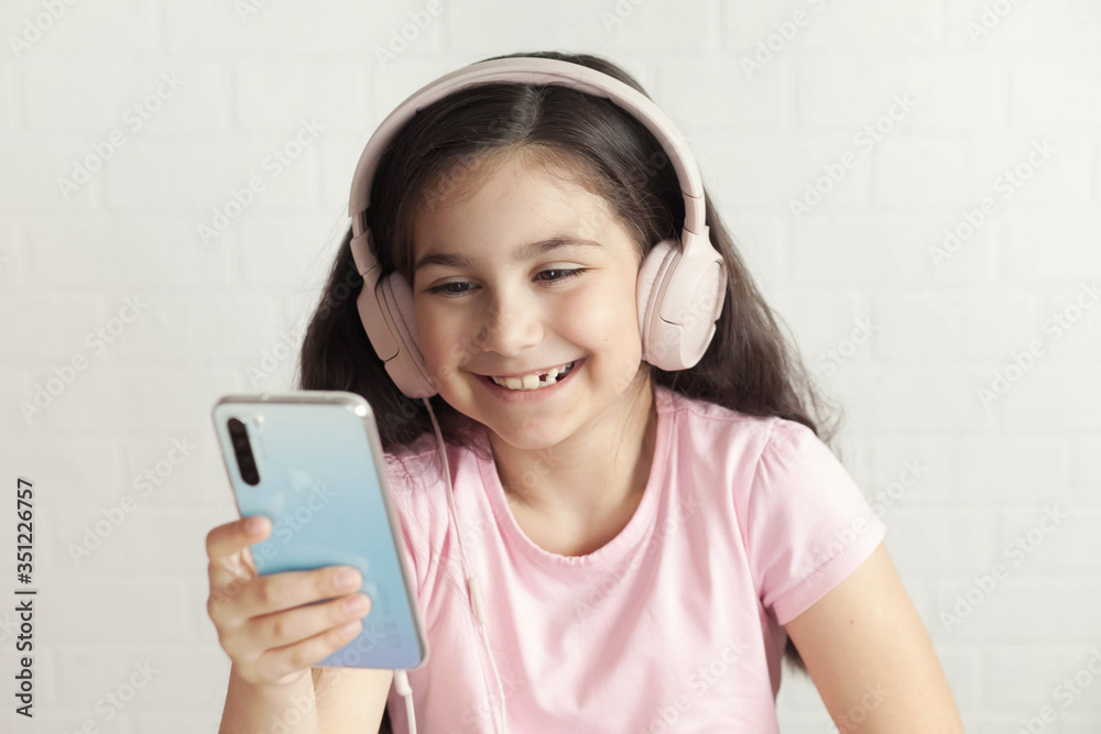 Cute child girl in headphones is using a smartphone, looking at camera and smiling on light background. Listening music. Online talking. Cute child girl in headphones is using a smartphone, looking at camera and smiling on light background. Listening music. Online talking.