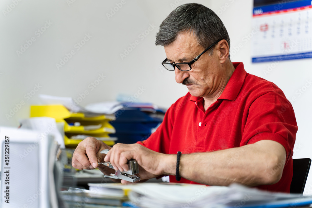 Side view of senior man caucasian mature entrepreneur salesman sitting by the desk at his shop wearing red shirt and eyeglasses repairing refill stapler holding in his hand in day on workplace Side view of senior man caucasian mature entrepreneur salesman sitting by the desk at his shop wearing red shirt and eyeglasses repairing refill stapler holding in his hand in day on workplace