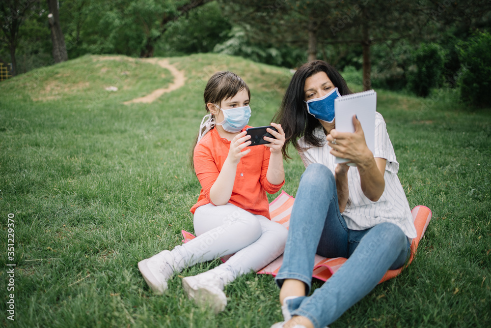 Little girl with protection mask looking at notebook in her mother's hands. Woman wearing antivirus mask while resting in park with her daughter who is holding phone. Little girl with protection mask looking at notebook in her mother's hands. Woman wearing antivirus mask while resting in park with her daughter who is holding phone.