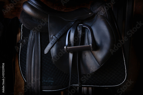 Fotografija Shiny black leather horse saddle in the dark stable , with stirrup, ready for ri