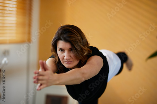 Middle aged caucasian yogi woman standing in warrior yoga pose at home.