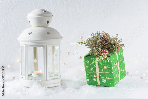 Merry Christmas Greeting Card. White retron lantern with burning candle and green gift box on white background covered by fresh snow photo