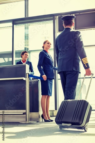 Portrait of young beautiful airport staff opening the gate for boarding while mature pilot walking in airport 