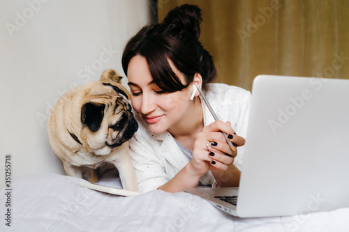 Young beatiful girl is smiling near a dog on bed while working at home in isolation. Home office. Concept of working in isolation. photo