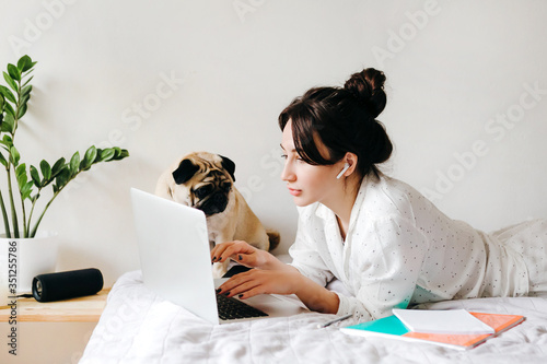 Young beatiful girl is working with laptop on bed near a dog at home in isolation. Home office. Concept of working and studying in isolation. photo