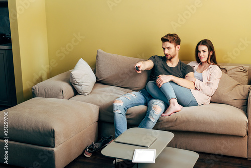 bearded man watching movie near girlfriend, laptop and digital tablet with white screen on coffee table