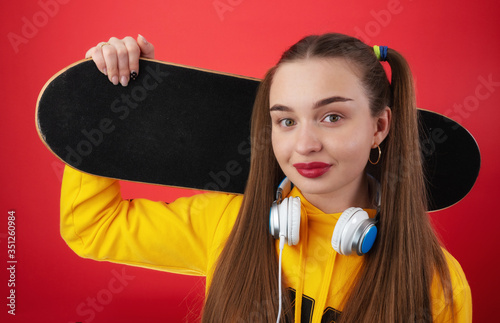 Modern young female in yellow hoodie with headphones on neck holding skateboard on shoulder and looking at camera against red background
