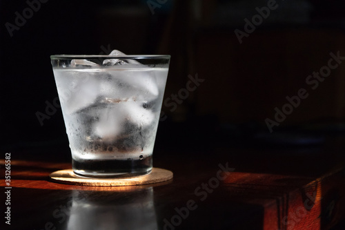 Glass of water with ice cubes on wooden table; shades of sunlight through from window.