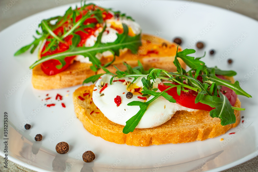 delicious poached eggs on crispy slices of wheat bread with slices of tomato, arugula, spices and black pepper on a white plate on a concrete background
