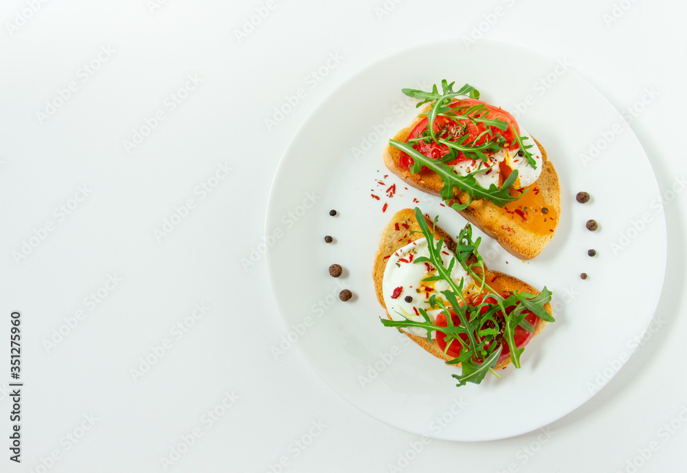 delicious poached eggs on crispy slices of wheat bread with slices of tomato, arugula, spices and black pepper on a white plate on a white background
