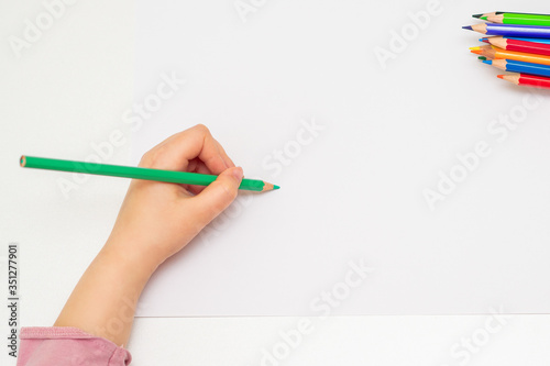 Top view of child's left hand drawing at blank white paper with a green pencil. Copy space for text. Mockup.