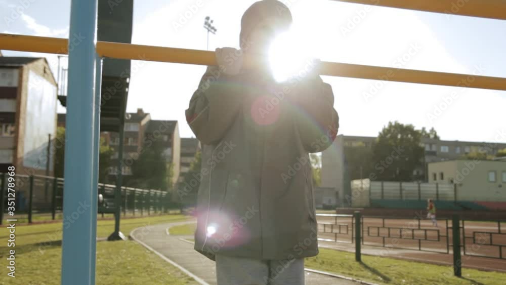 Boy doing pull ups on horizontal bar at stadium. Active kid in warm sportswear exercising in stadium and doing pull ups on metal sports equipment