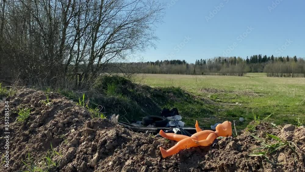 an orange children's toy lies on a hillock of land with green grass in the sun, followed by a spring field with a bunch of old garbage. bags, rags, broken glass, disposable tableware, canister. dump