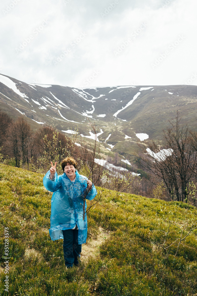 Happy smiling traveller senior beautiful woman in blue rain jacket and jeans in mountains surrounded by forest, enjoying silence and harmony of nature Happy smiling traveller senior beautiful woman in blue rain jacket and jeans in mountains surrounded by forest, enjoying silence and harmony of nature