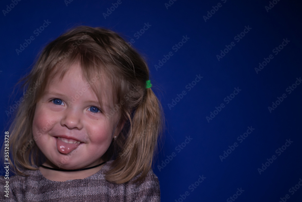 portrait of happy a three year old blond girl with blue eyes showing her tongue on a blue background portrait of happy a three year old blond girl with blue eyes showing her tongue on a blue background