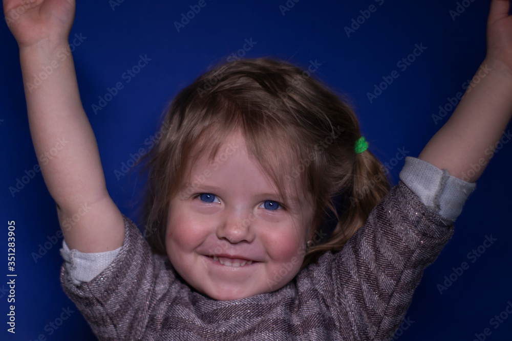 portrait of happy a three year old blond girl with blue eyes on a blue background - hands up