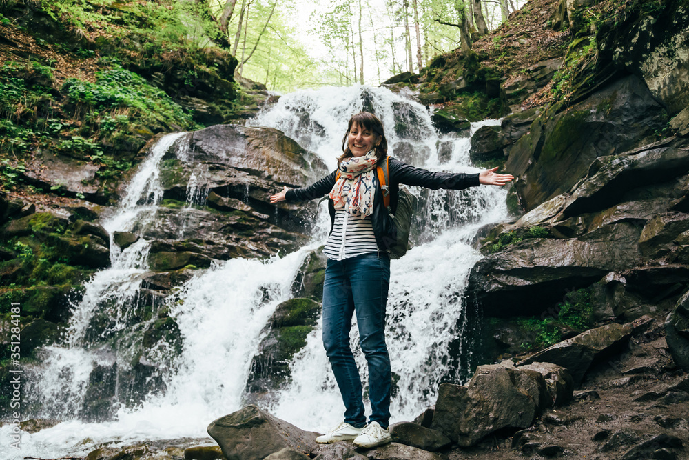 Happy smiling tourist girl in blue jeans, striped shirt and black jacket with backpack near to the cold fresh waterfall. Waterfall Shypit, cascade in Pylypets in the forest. Carpathian Mountains Happy smiling tourist girl in blue jeans, striped shirt and black jacket with backpack near to the cold fresh waterfall. Waterfall Shypit, cascade in Pylypets in the forest. Carpathian Mountains