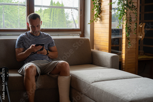 Man with broken leg in cast using tablet while sitting in sofa at home